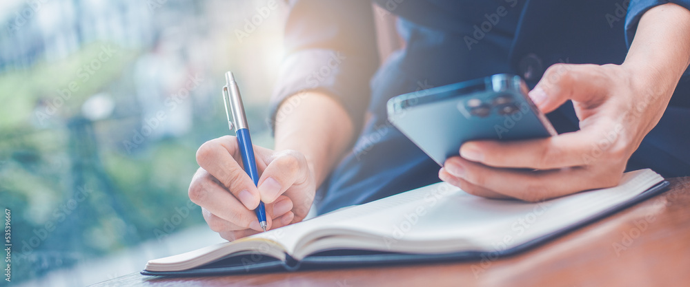 Woman hand writing on a notepad with a pen and she is holding a cell phone in the office.