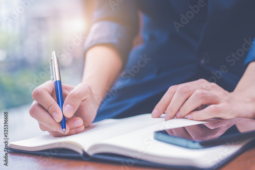 Obraz na płótnie Woman hand writing on a notepad with a pen and she is holding a cell phone in the office. w salonie