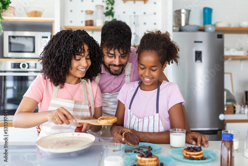 Glad positive young family foolishes with small daughter at kitchen, making pancakes, have tasty breakfast, pose at camera, drink fresh milk, eat pancakes © Graphicroyalty