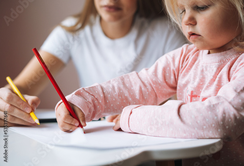 Little girl toddler with her mother drawing with colored pencils on table in children s room at home
