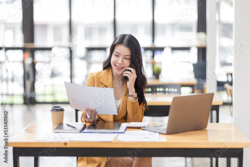 Asian Businesswoman using tablet notes while making phone call working laptop computer at workplease, Financial, accounting, report, tax, Business concept