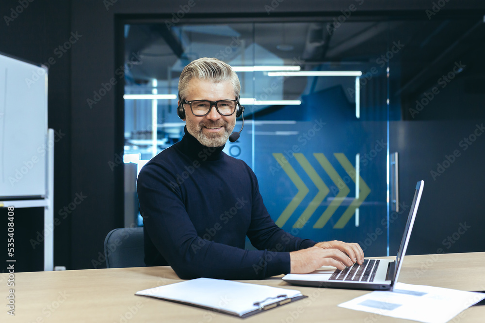 A gray-haired handsome businessman with a headset and a microphone is working on a laptop. Sits in the office at the table, conducts video conferences, consults. He looks at the camera, smiles. A gray-haired handsome businessman with a headset and a microphone is working on a laptop. Sits in the office at the table, conducts video conferences, consults. He looks at the camera, smiles.