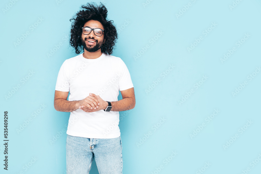 Handsome smiling hipster model.Sexy unshaven Arabian man dressed in white summer t-shirt and jeans clothes. Fashion male with long curly hairstyle posing near blue wall in studio. In glasses Handsome smiling hipster model.Sexy unshaven Arabian man dressed in white summer t-shirt and jeans clothes. Fashion male with long curly hairstyle posing near blue wall in studio. In glasses