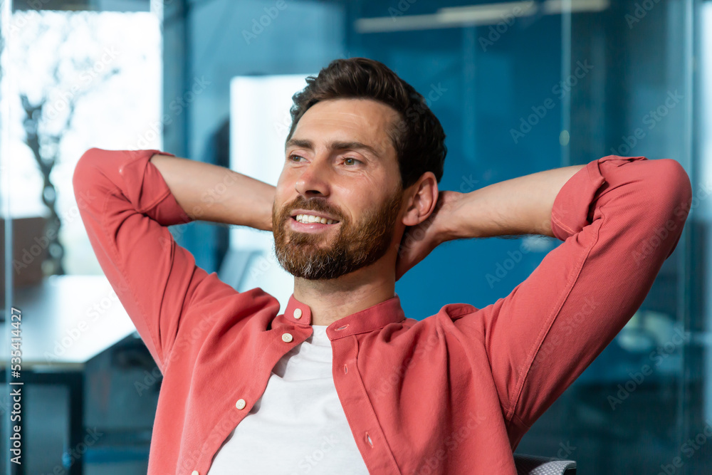 Young successful businessman startuper rests in office, man in red shirt throws hands behind head and dreams visualizes, designer thinks about new project. Young successful businessman startuper rests in office, man in red shirt throws hands behind head and dreams visualizes, designer thinks about new project.