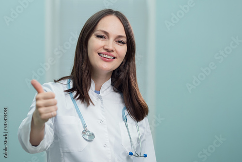Young woman pediatrician smiles looking straight ahead. Cheerful doctor shows patients OK gesture with hands. Lady prepares for appointment in ultrasound office
