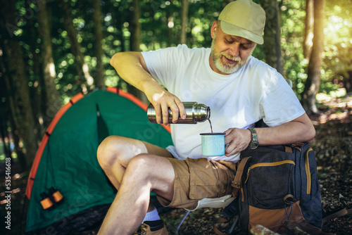 Portrait of mature man sitting in the forest with a tent in background and pours coffee from a thermos.