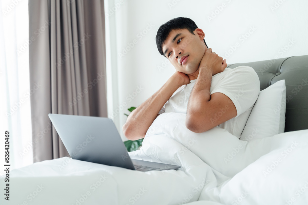 Asian man in white t-shirt laying on bed using laptop computer having neck pain. Asian man in white t-shirt laying on bed using laptop computer having neck pain.