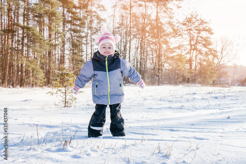 Little girl 3-4 years old in winter clothes overalls, hat and mittens jumps on snow and laughs against snow-covered pines and fir trees on sunny day. Winter holidays, children playing outdoors
