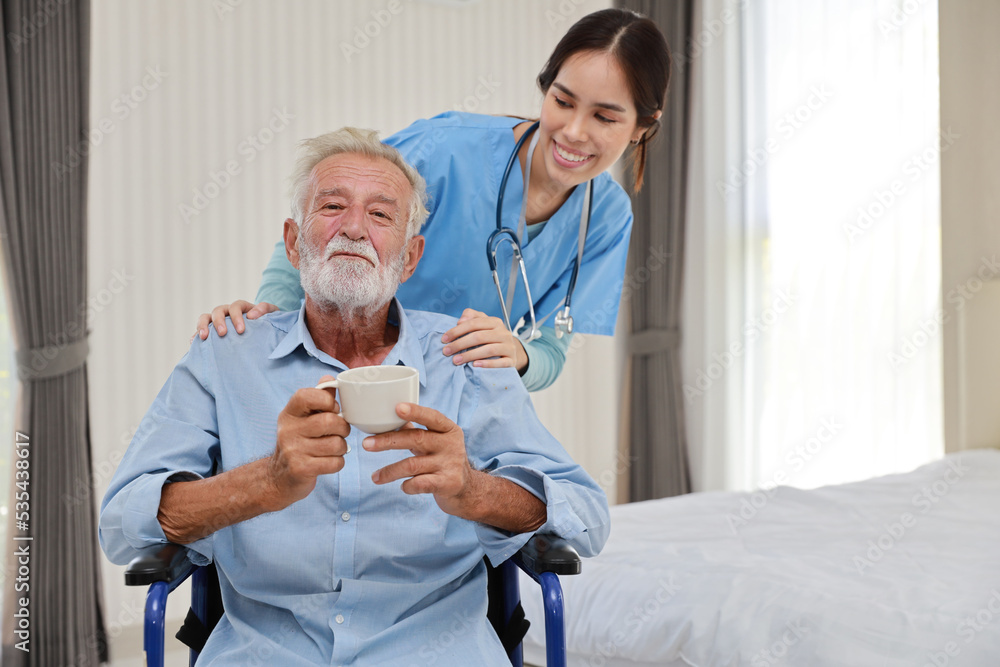 Fototapeta premium Happy caregiver giving senior caucasian man with wheelchair a cup of coffee or tea for breakfast in bedroom at retirement house. Asian smiling nurse taking good help care and support elder patient.