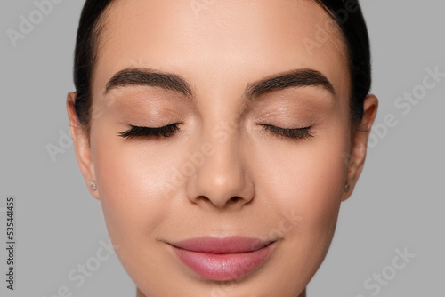 Beautiful young woman showing extended and ordinary eyelashes on grey background, closeup