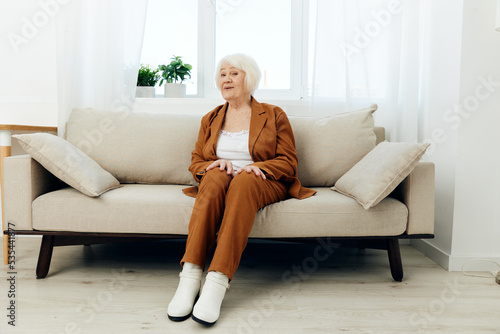 a sweet, amiable elderly woman in a brown suit is sitting on a beige wide sofa, smiling pleasantly while in the comfortable environment of her apartment