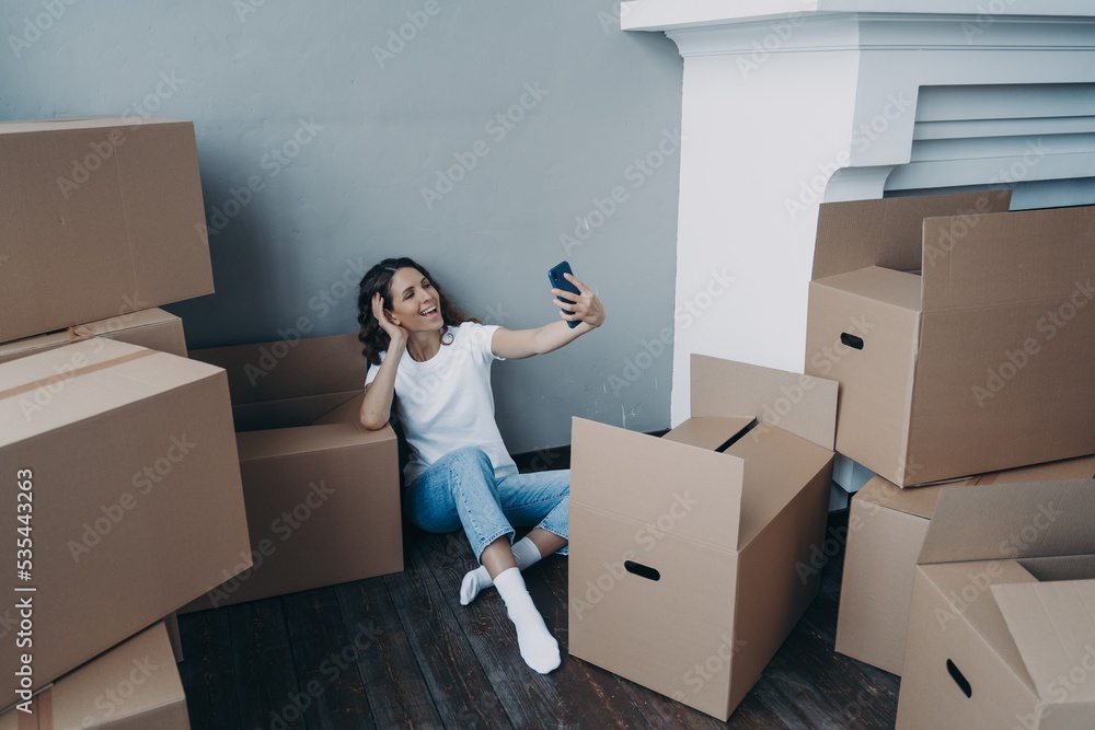 Joyful girl chatting by video call sitting with cardboard boxes during packing things for relocation Joyful girl chatting by video call sitting with cardboard boxes during packing things for relocation