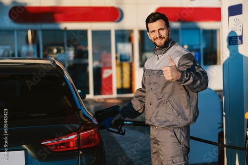 A gas station worker fills up the car tank and gives thumbs up while smiling at the camera.