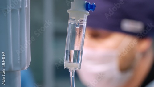 Freeze frame of a falling drop in a drip system with medicines. In the background, the concentrated face of a surgeon in a mask and surgical clothes can be seen.
