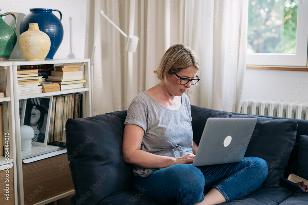 mid adult woman sits on sofa in living room and using laptop computer mid adult woman sits on sofa in living room and using laptop computer
