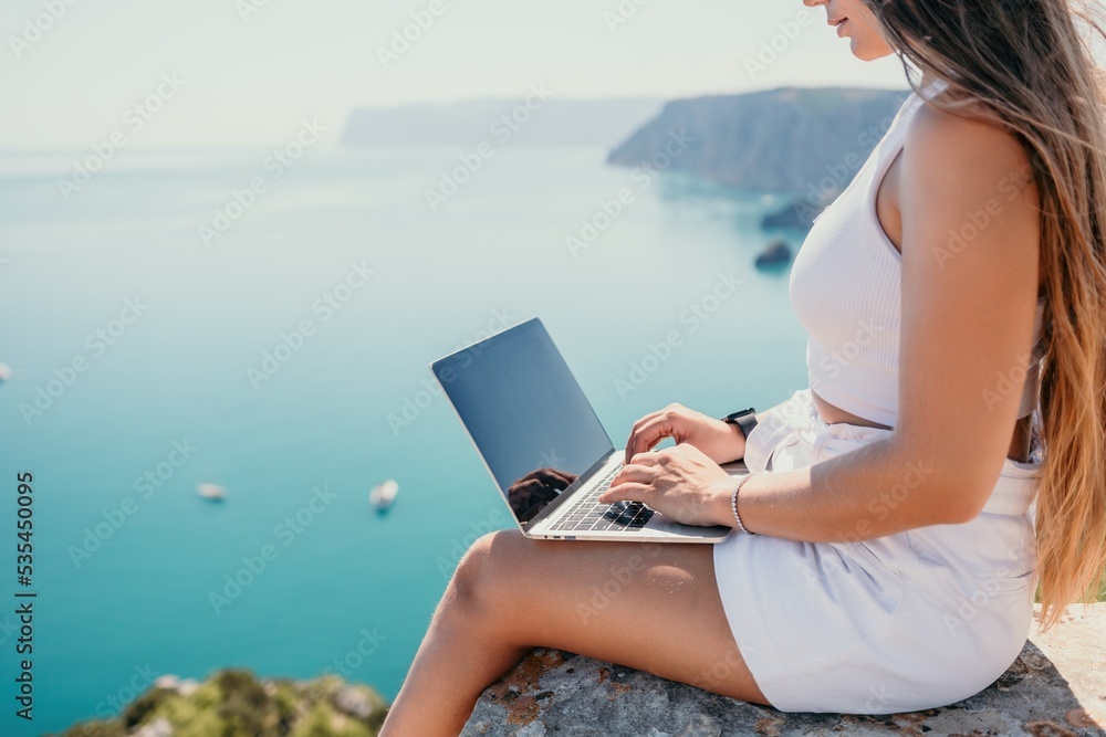 Naklejka premium Digital nomad, woman in the hat, a business woman with a laptop sits on the rocks by the sea during sunset, makes a business transaction online from a distance. Freelance, remote work on vacation.