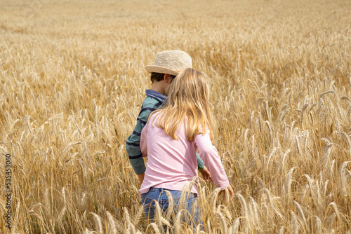 Golden wheat field an two kids, twins, a blonde girl with pink shirt and jeans, a boy with a hat, and a green and blue shirt, both playing and bonding together © Julia