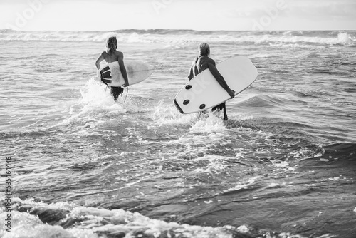 Father and son running on the beach at sunset for surf training - Focus on senior man holding longboard - Black and white editing