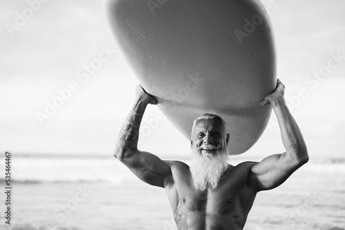 Happy senior surfer man holding surf board on the beach at sunset - Focus on face - Black and white editing