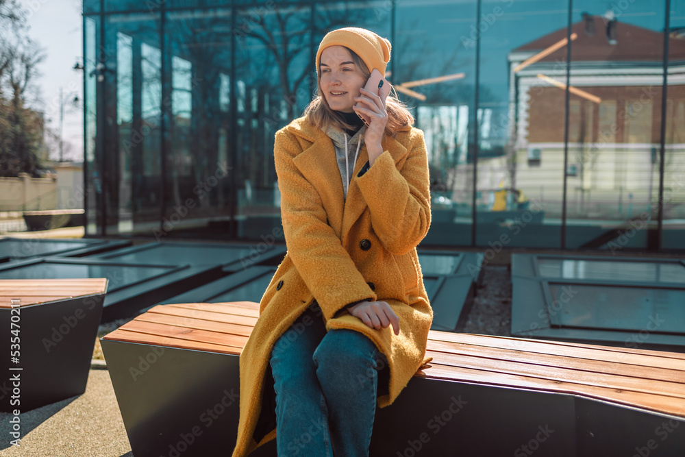 Fototapeta premium Young smiling caucasian fun woman in bright clothes sits on a stylish wooden bench in the city museum outdoors use mobile cell phone chat with boyfriend on social network. People active urban