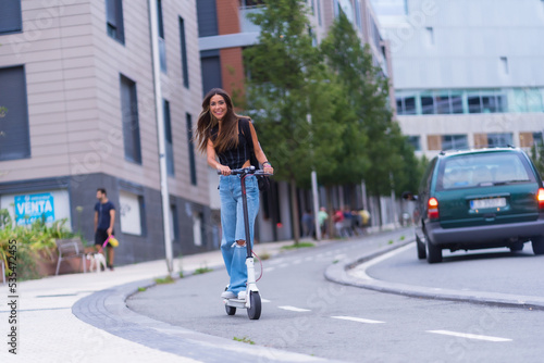 A young brunette woman moving in the city with an electric scooter smiling, along the bike path © unai