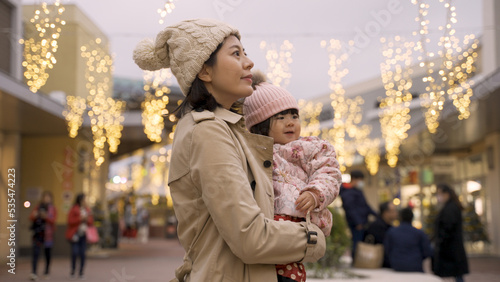 smiling asian mother and baby turning and looking around at beautiful outdoor Christmas lights above head at a mall while shopping for the winter holiday