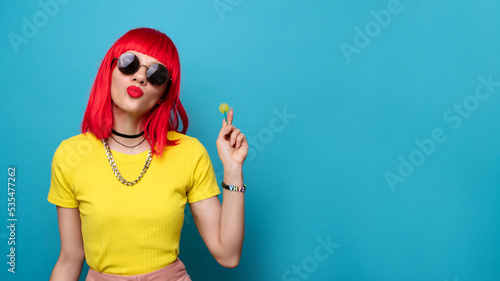A bright and funny young woman with red hair licks a lollipop. Studio colorful picture of a pin-up style woman with a lollipop on a stick, on a blue background. A place for your text.