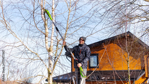 Electric chain pruner with a telescopic pole close up. Winter tree pruning. A man gardener standing on a stepladder cuts branches on a tree with a special electric saw.