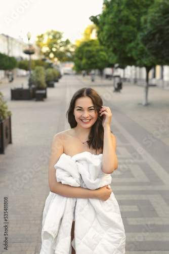 Beautiful young woman wrapped in a white blanket on an empty city road in the morning at dawn. © Artem Zakharov