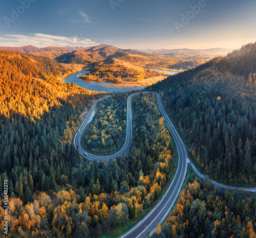 Aerial view of mountain road in colorful forest at sunset in autumn in Ukraine. Top view from drone of road in woods in fall. Beautiful landscape with highway in hills, river, pine trees, mountains © den-belitsky
