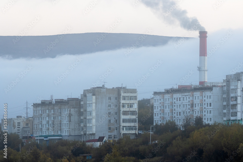 View of the buildings of a residential city block and a smoking chimney of a coal-fired combined heat and power plant. Foggy weather. Ecology in the cities of Siberia. Magadan, Magadan region, Russia.