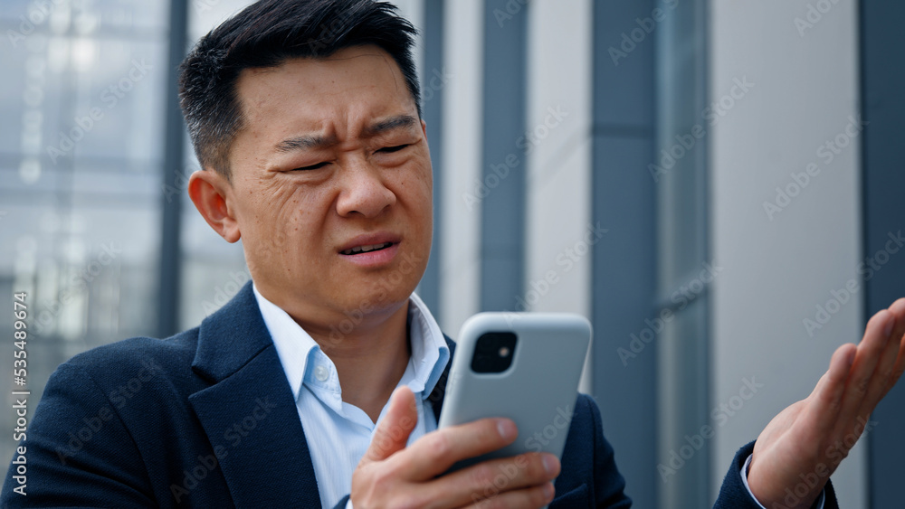 Close up Asian middle-aged adult man mad unhappy businessman feeling annoyed with using broken smart phone low battery problem angry having problem with mobile telephone frustrated bad news outdoors