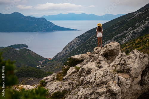 a beautiful girl in a white dress stands on a hill gazing at the panorama of the peljesac peninsula in croatia, mighty mountains and cliffs over the mediterranean sea