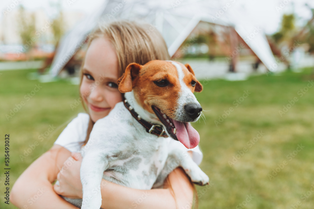 a little girl kisses and hugs her Jack Russell terrier dog in the park. Love between the owner and the dog. a child is holding a dog in his arms. a little girl kisses and hugs her Jack Russell terrier dog in the park. Love between the owner and the dog. a child is holding a dog in his arms.