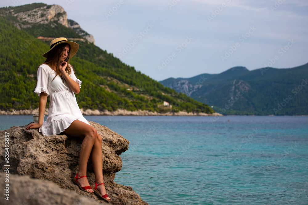 beautiful tanned woman in white dress and hat rests on the rocks during sunset over paradise beach in croatia with green mountains in the background; peljesac peninsula 