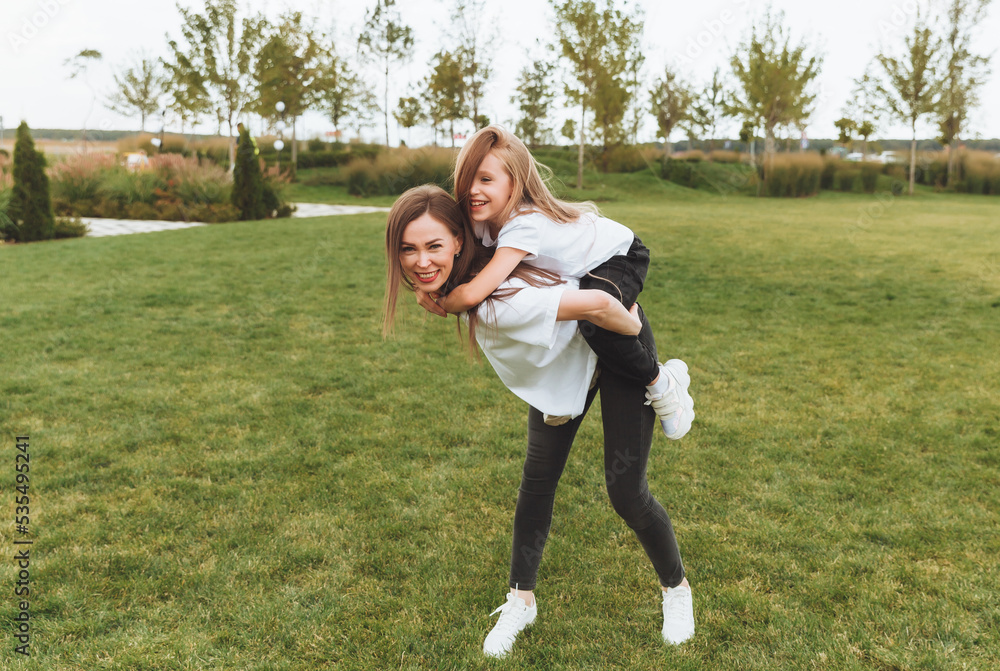 A happy mother and a little girl play and have fun in the park together, a girl rides on her mother's back. people in white t-shirts