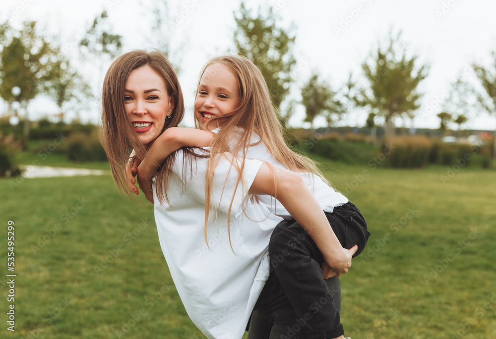 A happy mother and a little girl play and have fun in the park together, a girl rides on her mother's back. people in white t-shirts