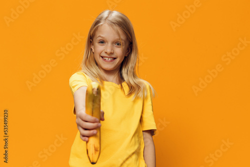 cute, shy, beautiful school-age girl in yellow clothes stands against a bright, yellow background, holding out a banana to the camera. Horizontal photo with empty space for inserting advertising text