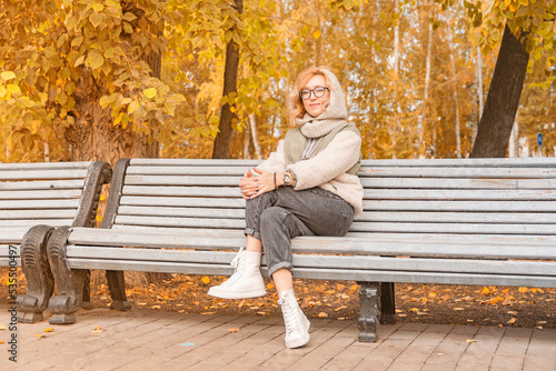 Young and beautiful blonde girl in glasses and insulated clothes sits on bench in autumn recreation park against background of yellowing trees. Looks at camera.