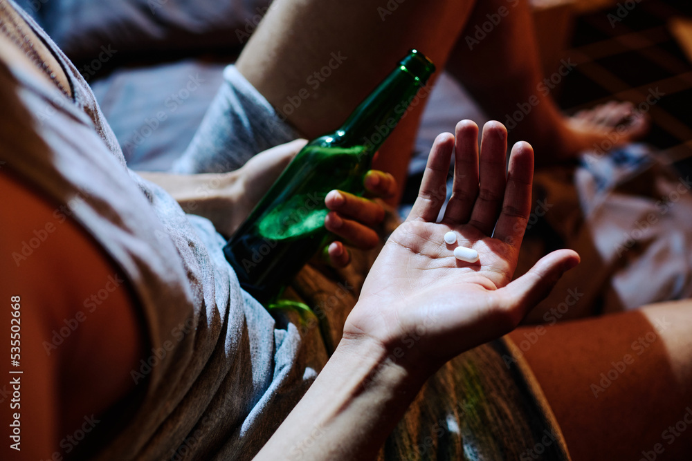 Above angle of hand of young depressed man holding sleeping pills or antidepressants and bottle of alcohol while sitting on bed
