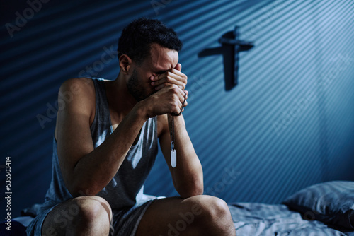 Young depressed man with two medallions on silver chain crying or grieving about loss of someone he loved while sitting on bed at night
