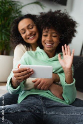 happy african american preteen girl waving hand near curly mother during video call on smartphone.