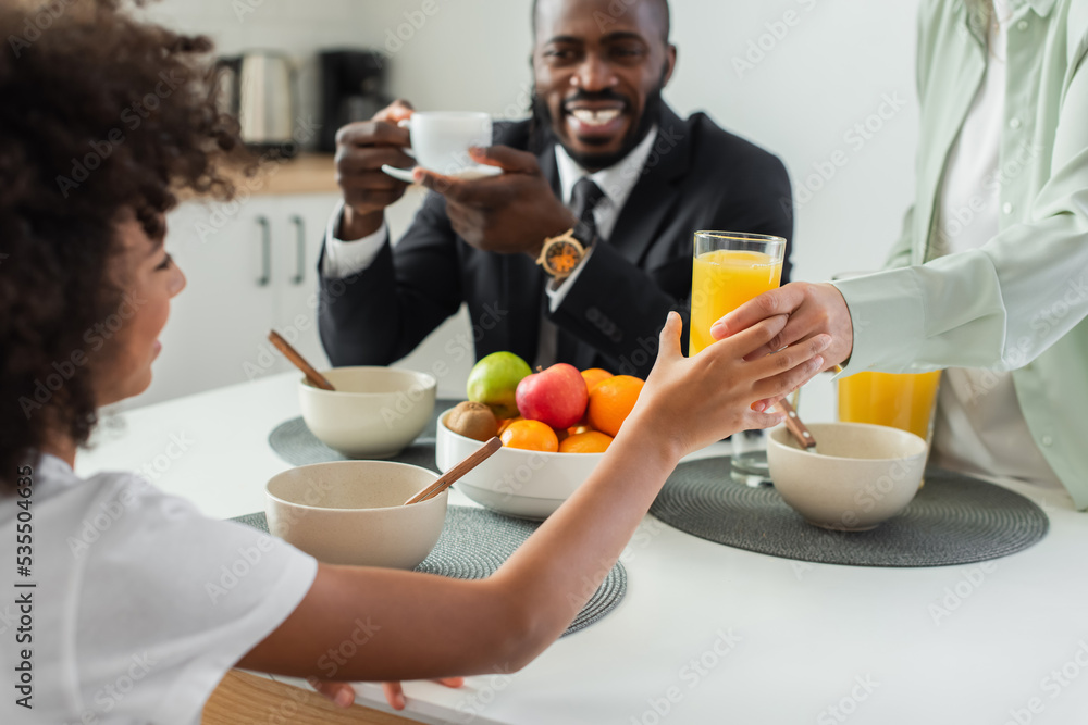 african american woman giving glass of orange juice to happy daughter near husband during breakfast. african american woman giving glass of orange juice to happy daughter near husband during breakfast.