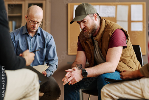 Young upset man describing traumatic event or experience from his life while sitting among counselor and other attendants of session