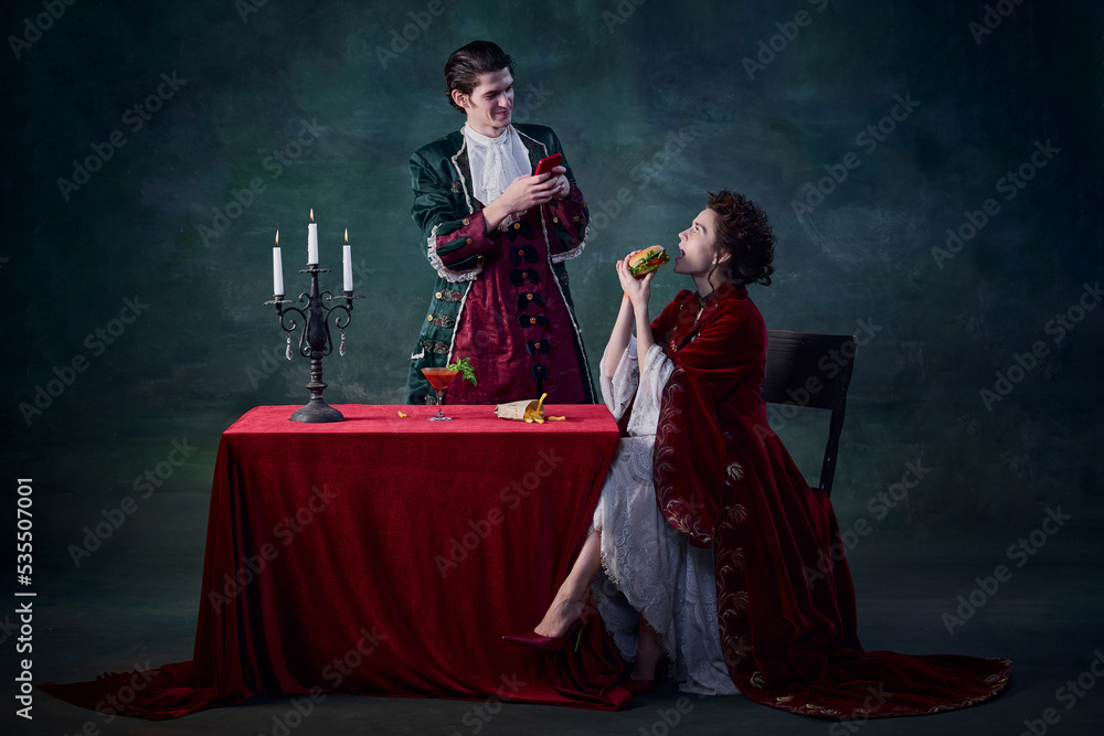 Fototapeta premium Portrait of man and woman in image of vampires over dark green background. Man taking selfie of woman eating burger