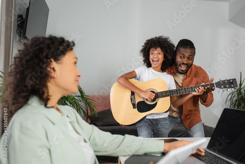curly african american girl playing acoustic guitar near father and mother working from home.