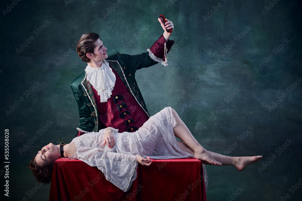 Portrait of man in image of medieval vampire taking selfie of dead woman lying on table over dark green background. Victim Portrait of man in image of medieval vampire taking selfie of dead woman lying on table over dark green background. Victim