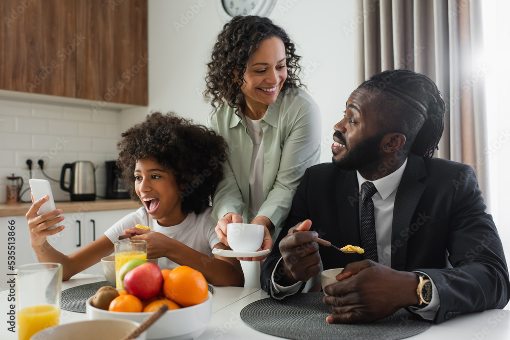 cheerful african american woman holding cup of coffee near husband in suit while preteen daughter using smartphone. cheerful african american woman holding cup of coffee near husband in suit while preteen daughter using smartphone.