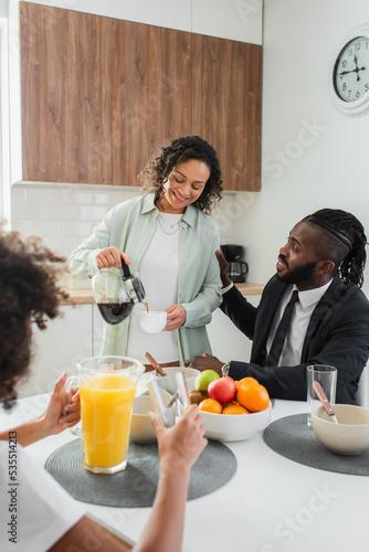 happy african american woman pouring coffee in cup near husband in suit and preteen daughter during breakfast.