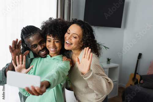 cheerful african american family smiling and waving hands during video call on smartphone.